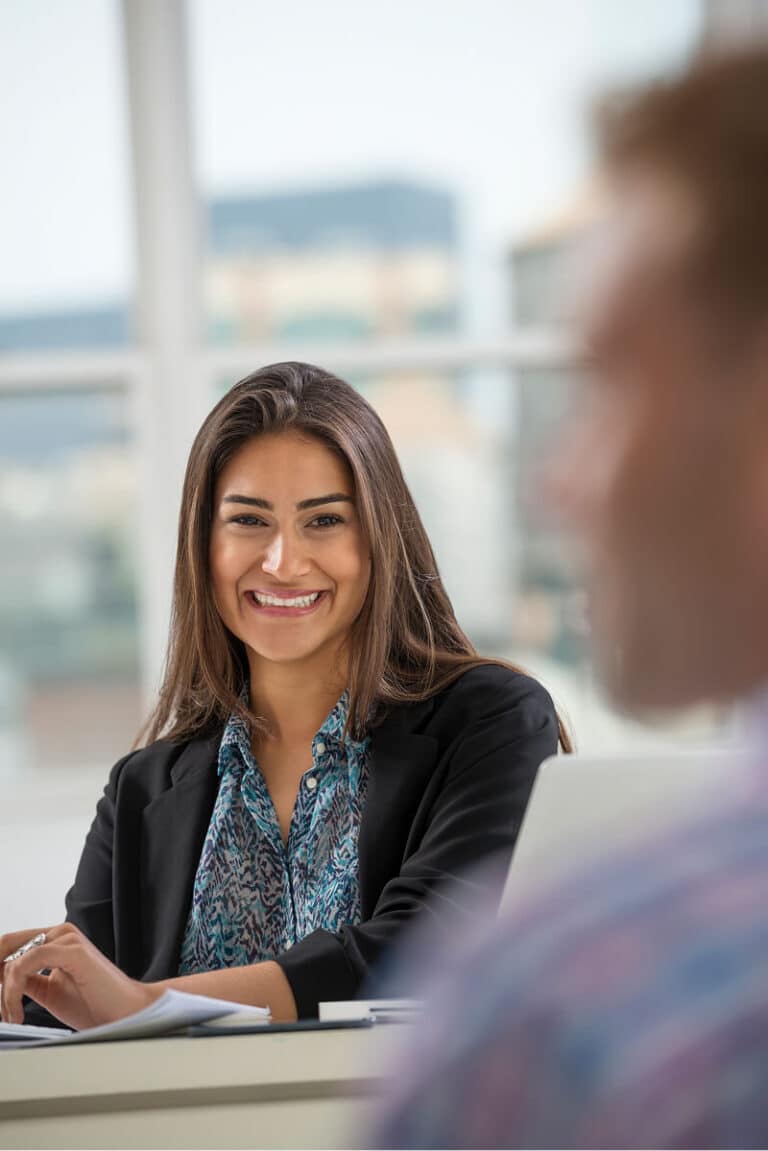 A woman in business attire sits at a desk and smiles, looking toward a person in the foreground during a meeting in a bright office setting.