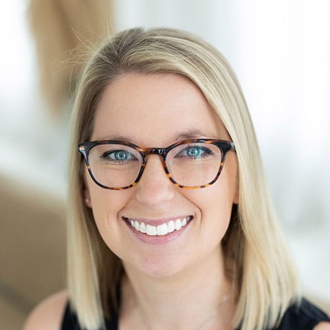 A woman with straight blonde hair, wearing tortoiseshell glasses and a black top, smiling at the camera with a blurred light background.