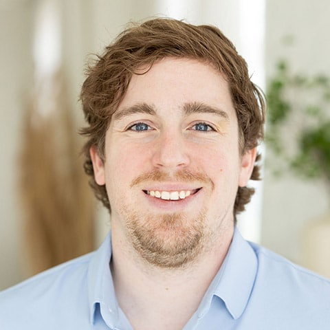 A man with light skin, brown hair, and a short beard is smiling at the camera, wearing a light blue collared shirt. The background is softly blurred.