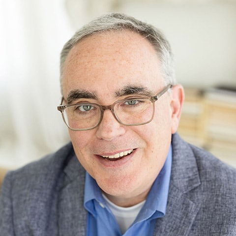 A middle-aged man with gray hair and glasses, wearing a blue shirt and gray blazer, smiling at the camera in a well-lit indoor setting.