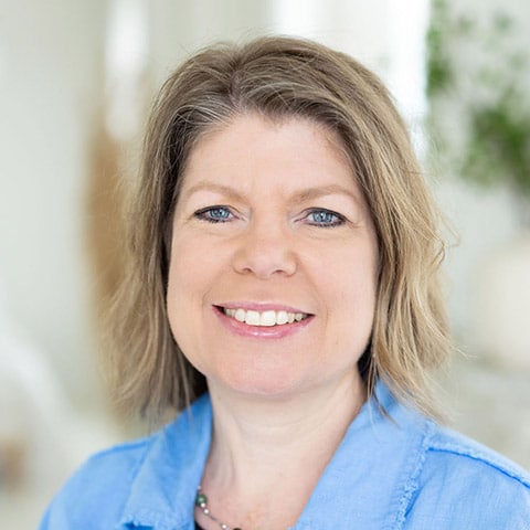A woman with light brown hair and blue eyes smiles at the camera. She is wearing a light blue shirt and a beaded necklace. The background is softly blurred.
