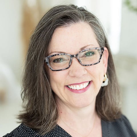 Woman with shoulder-length brown hair and glasses, wearing a black top with white dots, smiling at the camera in a well-lit room.