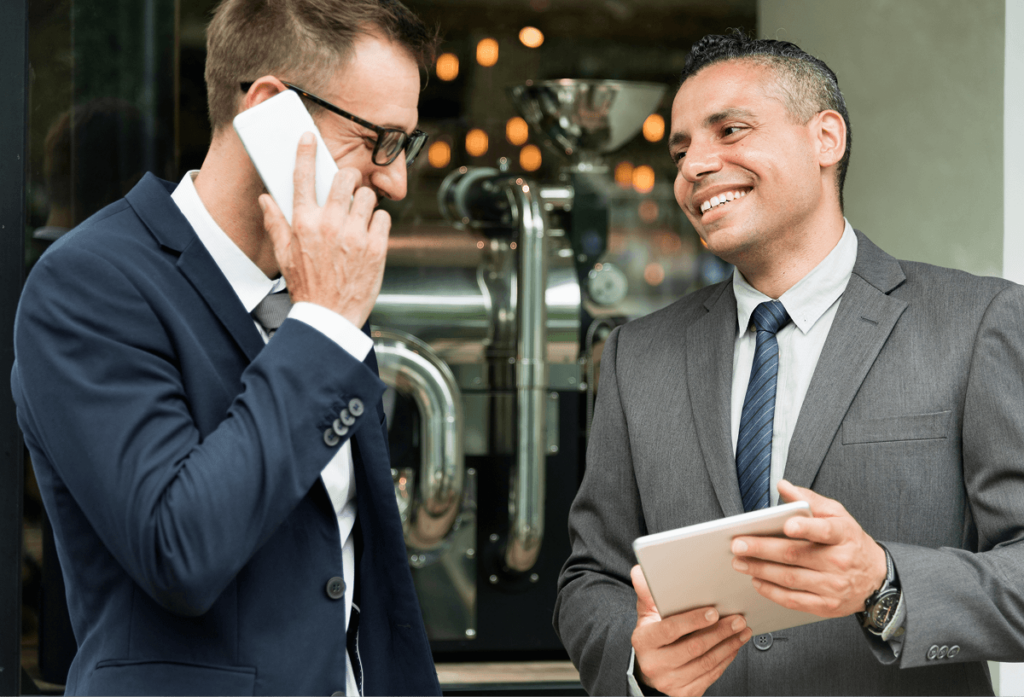 Two men in business suits stand outdoors; one is talking on a smartphone, and the other is smiling while holding a tablet. Industrial equipment is visible in the background.