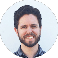 A man with dark hair and a beard is smiling at the camera. He is wearing a checked shirt and standing in front of a plain white background.