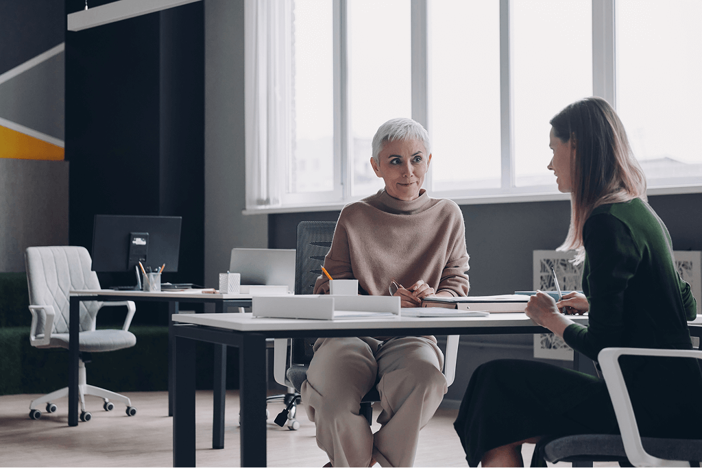 Two women sit across from each other at a desk in a modern office, engaged in conversation, with computers and office chairs visible in the background.