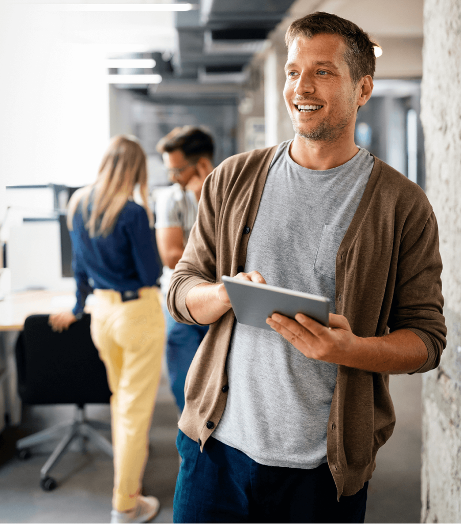 Smiling man holding a tablet stands in a modern office, while two people discuss work at a nearby desk in the background.