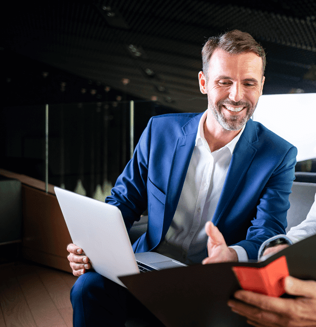 A man in a blue suit smiles while holding a laptop and looking at another person who is showing him a red smartphone in an office setting.