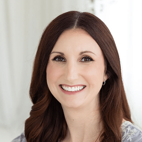 A woman with long brown hair and fair skin smiles at the camera against a softly lit, white background.