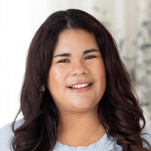 A woman with long, dark wavy hair smiles at the camera. She is wearing a light blue top and a necklace, with a soft-focus background.